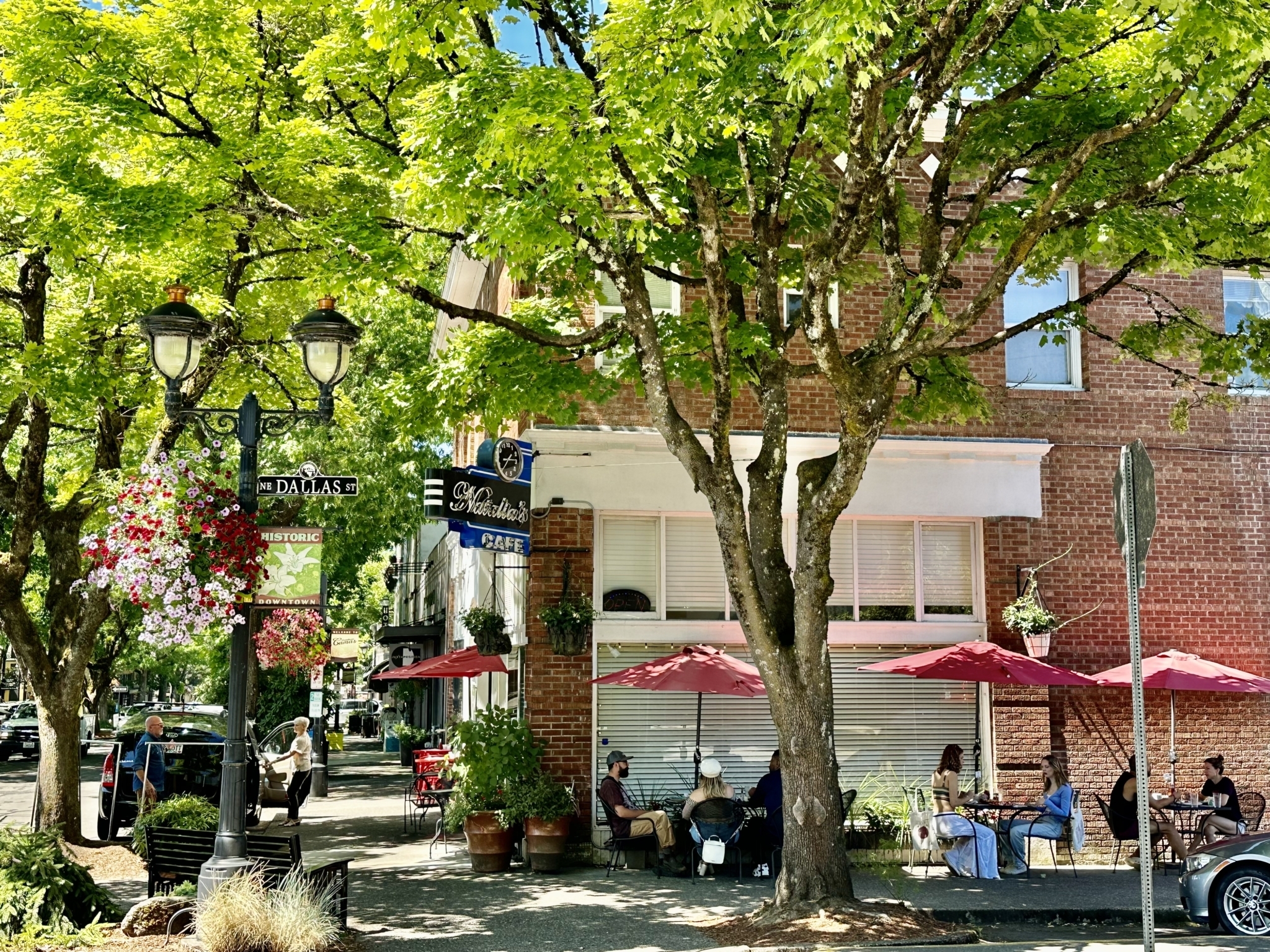 On a sunny day, diners enjoy outdoor sidewalk seating under the shade of trees and meals from a café located in a historic brick building.